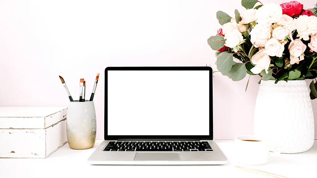 Ordinateur portable sur bureau blanc avec pinceaux, boîte de rangement, tasse et bouquet de roses dans un vase