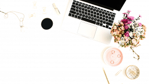 Ordinateur portable sur bureau blanc avec fleurs, café, accessoires dorés, lunettes et bougie rose