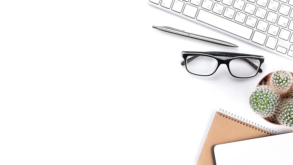 Clavier blanc, stylo argenté, lunettes noires, carnet kraft et cactus sur un bureau blanc