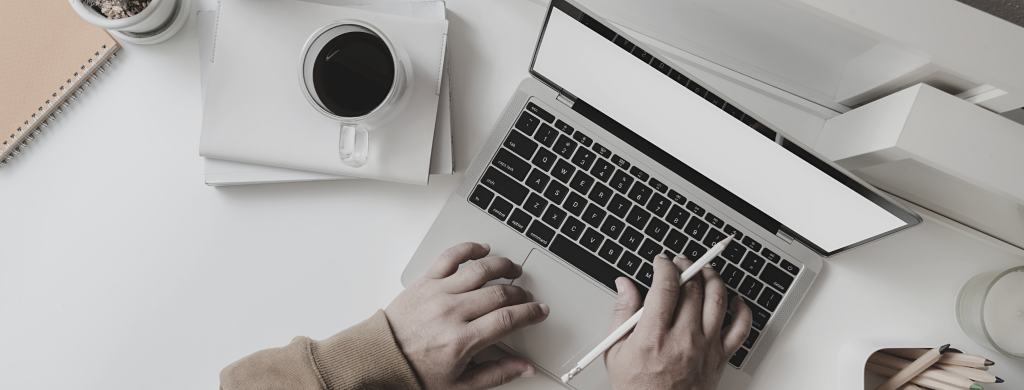 Personne utilisant un ordinateur portable avec un stylo à la main, tasse de café, carnet et écouteurs sur un bureau blanc